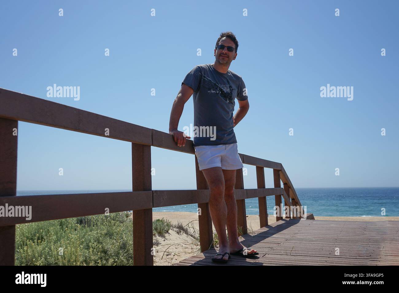 Un uomo europeo magro e dai capelli scuri, con una maglietta blu, pantaloncini bianchi e infradito, si rilassa su una passerella di legno in riva al mare. Foto Stock