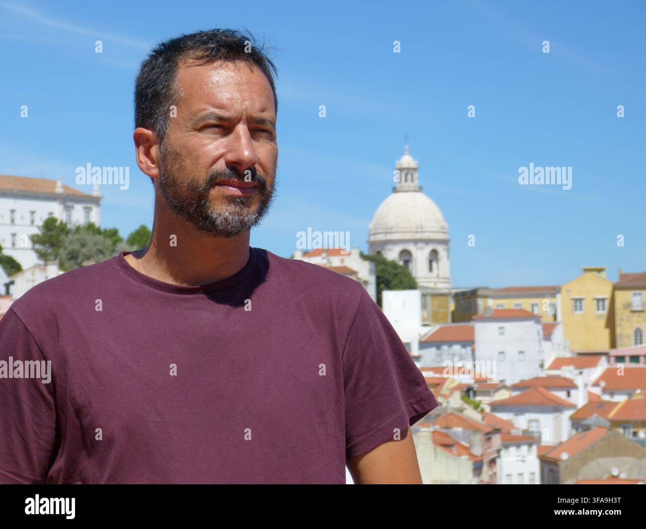 Un uomo europeo magro, dai capelli scuri e con la barba rasata, indossa una maglietta bordeaux e si rilassa a Lisbona, Portogallo. Foto Stock