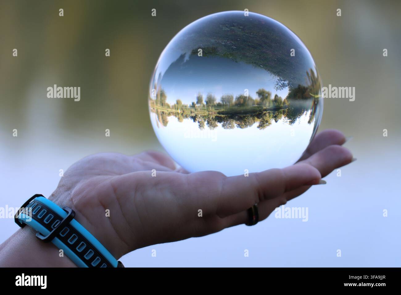 Primo piano di una mano femminile che tiene una sfera di lente cristallina che riflette un lago, con uno smartwatch, su uno sfondo sfocato morbido a bokeh Foto Stock