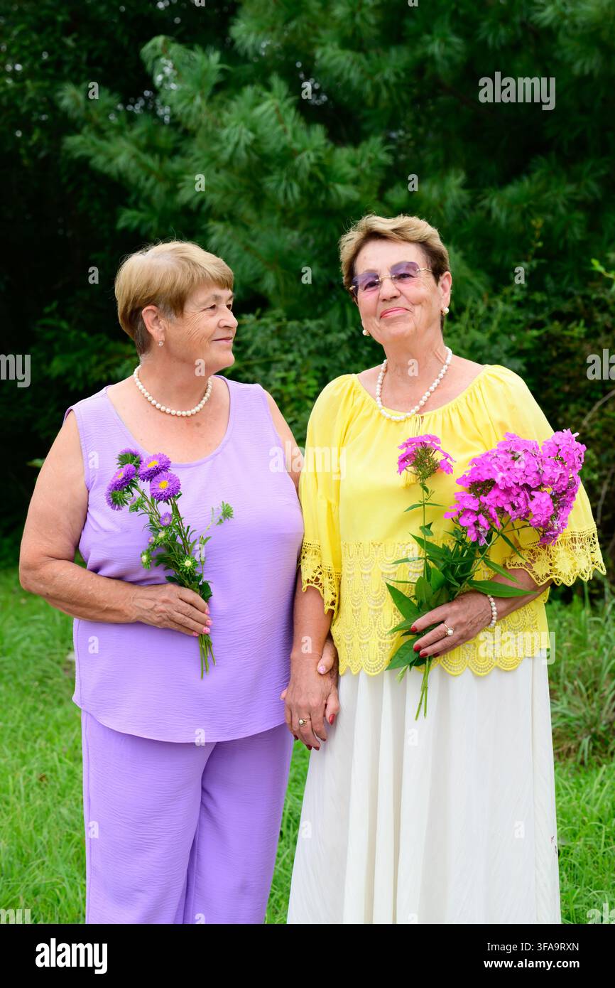 Due sorelle russe di 73 e 79 anni che tengono mazzi di fiori sorridendo e in piedi insieme in un giardino di un villaggio rurale. Foto Stock
