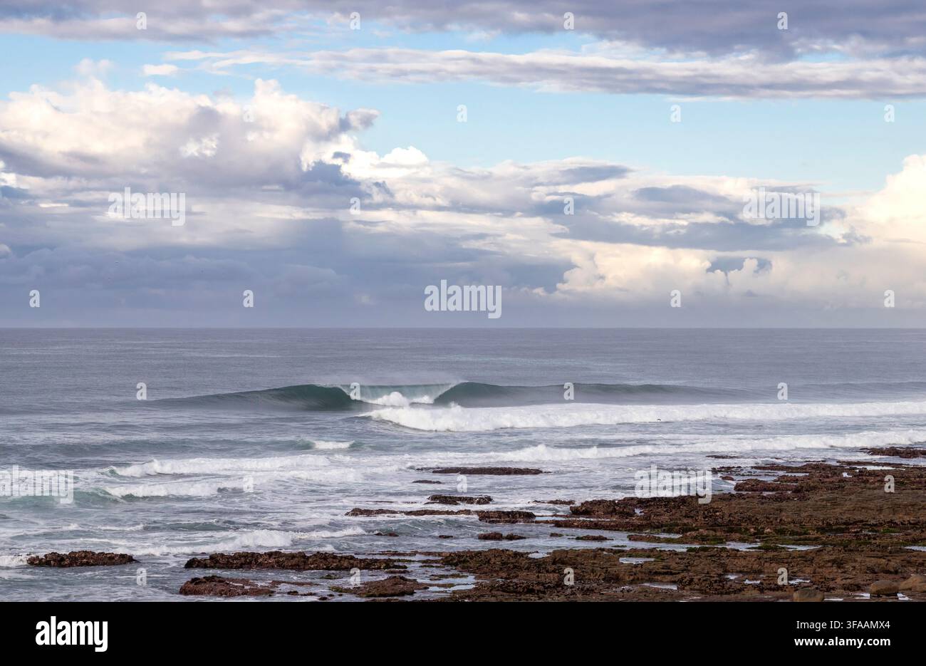Scena della costa del Capo Orientale con una riva rocciosa, onde morbide su un mare calmo sotto un cielo nuvoloso Foto Stock
