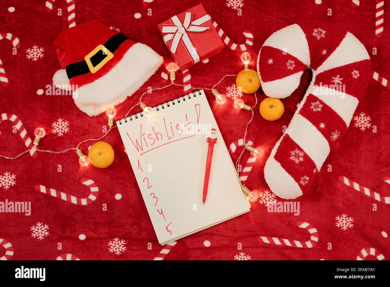 Composizione piatta festiva con una pagina vuota della lista dei desideri, cappello di Babbo Natale, penna rossa, scatola regalo, cuscino a forma di bastoncino di zucchero, mandarini e luci natalizie luminose su rosso Foto Stock