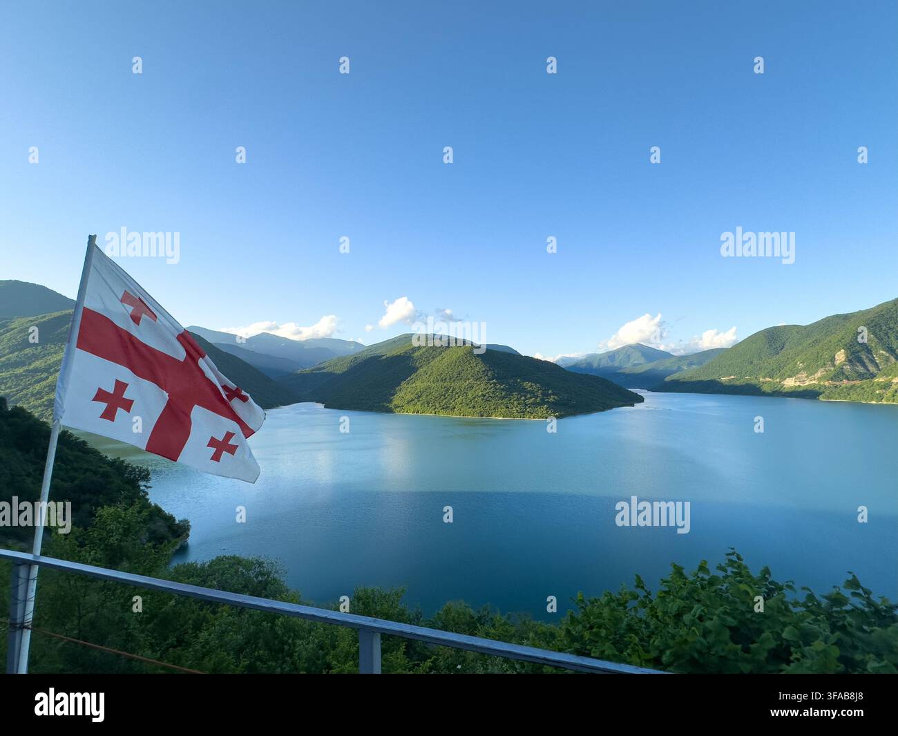 Bandiera georgiana che sventola sopra il bacino idrico di Zhinvali e le montagne verdi. Vista scenografica di una bandiera nazionale nel vento e del pittoresco Zhinvali, colline rigogliose, giornata soleggiata - Immagine stock catturata con smartphone