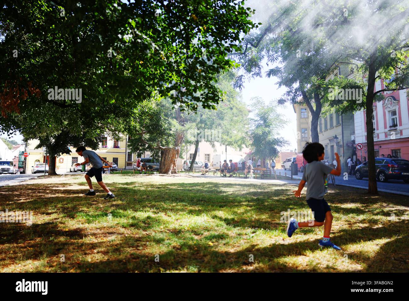 Praga, Repubblica Ceca - 8.14.2026: Due ragazzi che corrono e giocano all'aperto in un parco cittadino verde sotto una nebbia d'acqua in una calda giornata estiva. Foto Stock