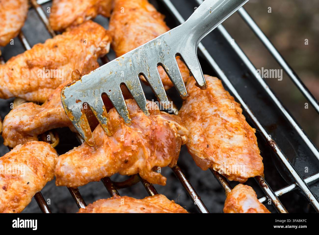 Prima vista di ali di pollo crude condite maneggiate con pinze metalliche su una griglia in metallo nera durante la preparazione del cibo all'aperto. Foto Stock