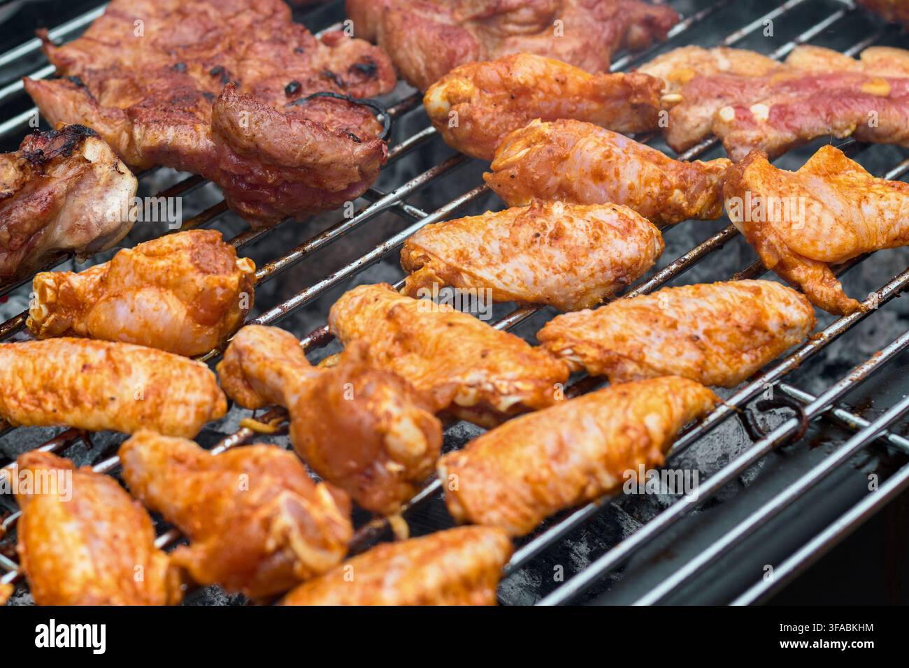 Deliziose ali di pollo condite e carne di maiale che cuociono su una griglia metallica calda sopra brace di carbone ardente durante una preparazione di pasti estivi all'aperto Foto Stock