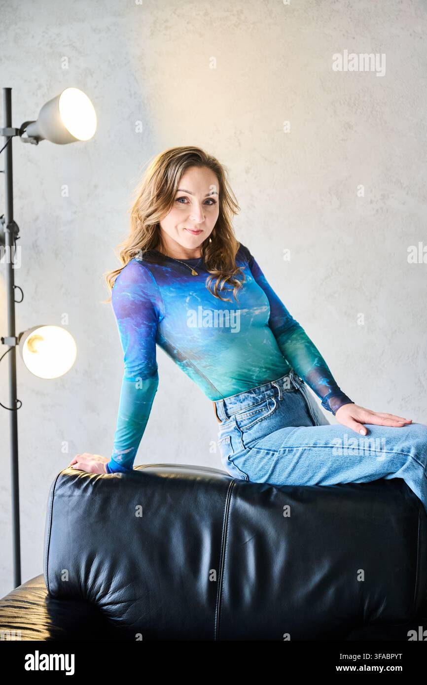 Donna seduta sullo schienale di un divano in pelle e che guarda la telecamera all'interno. Giovane donna adulta caucasica con capelli castani ondulati, top in mesh blu colorato, jeans di denim, atmosfera elegante e sicura di sé Foto Stock
