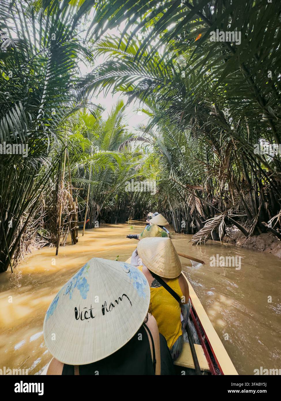 Vietnam - 17 agosto 2026: Vista di persone che indossano cappelli conici tradizionali mentre navigano su una barca di legno attraverso un canale stretto fiancheggiato da palme lussureggianti Foto Stock