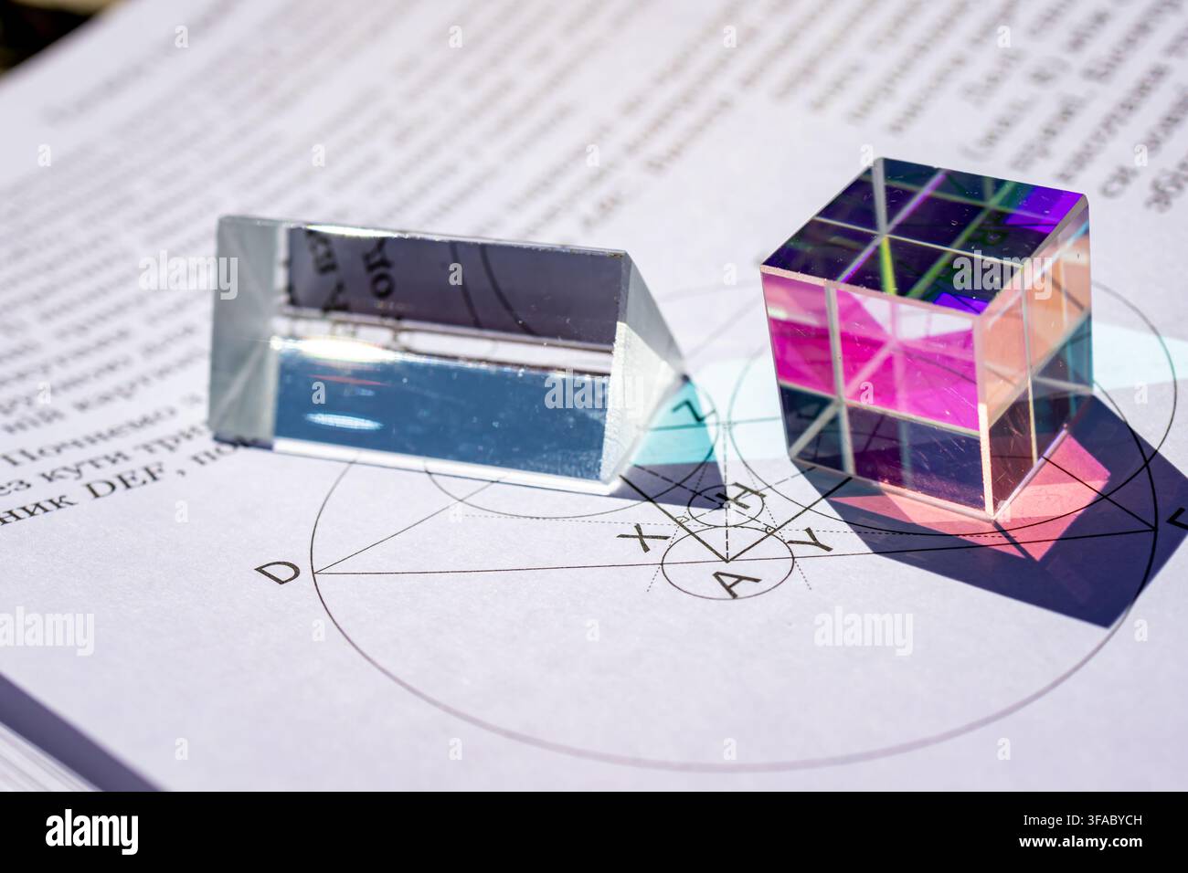 Prisma di vetro ottico e prisma cubo colorato appoggiati su un foglio di lavoro di geometria con una costruzione precisa del cerchio, la luce del sole proietta riflessi vividi rappr Foto Stock