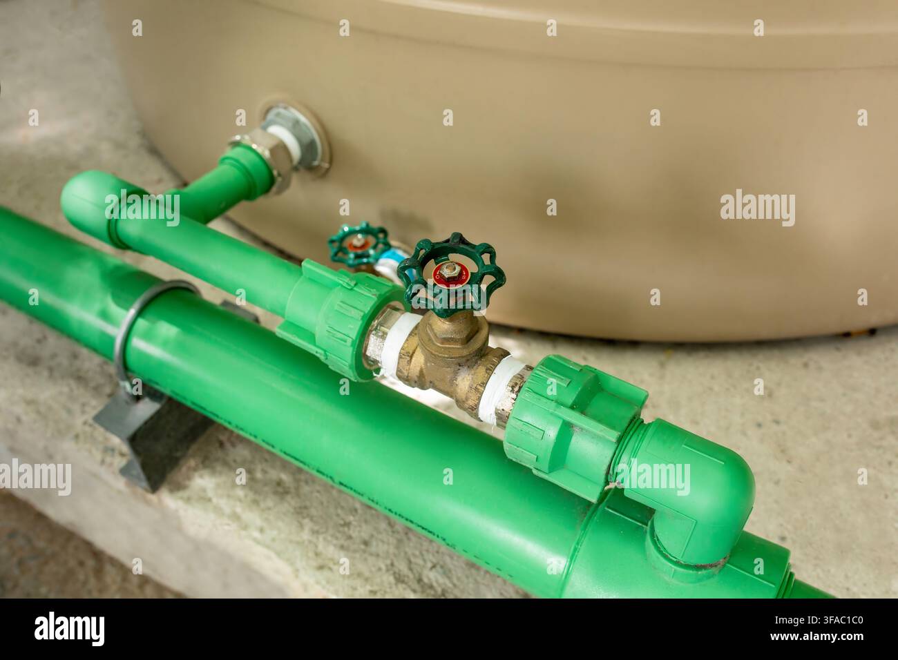 Primo piano di una ruota a valvola verde montata su un raccordo per tubi vicino a un serbatoio d'acqua beige, dettaglio dell'infrastruttura idraulica Foto Stock