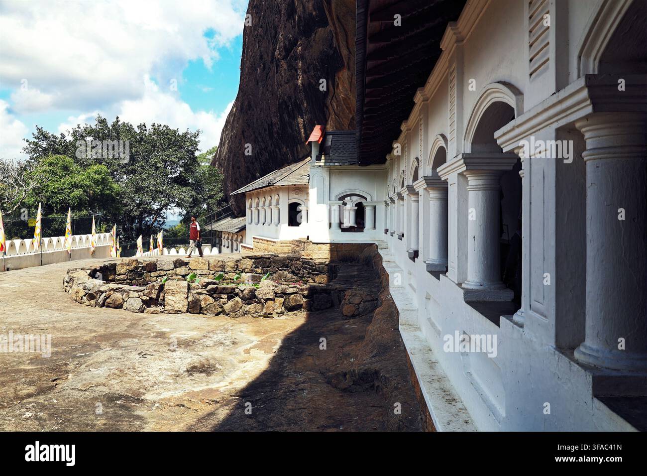 Uomo che passa davanti alla facciata colonnata bianca del tempio delle grotte storico di Dambulla, costruito sotto un enorme sporgione roccioso nello Sri Lanka Foto Stock