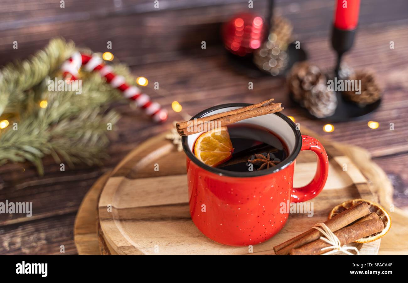 Vino caldo servito in una tazza di smalto rossa su un tagliere di legno con bastoncini di cannella, arance essiccate, rami di sempreverdi e decorazioni natalizie festive Foto Stock