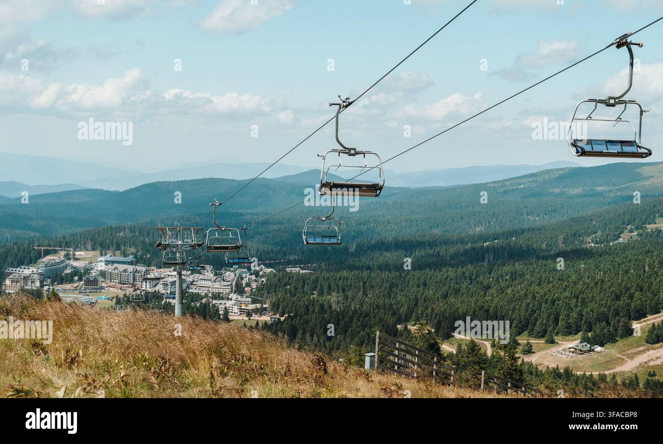 I sedili vuoti della seggiovia si estendono sopra la stazione montana di Kopaonik in Serbia. Pendici boschive ed edifici alberghi si estendono attraverso il paesaggio estivo. Foto Stock
