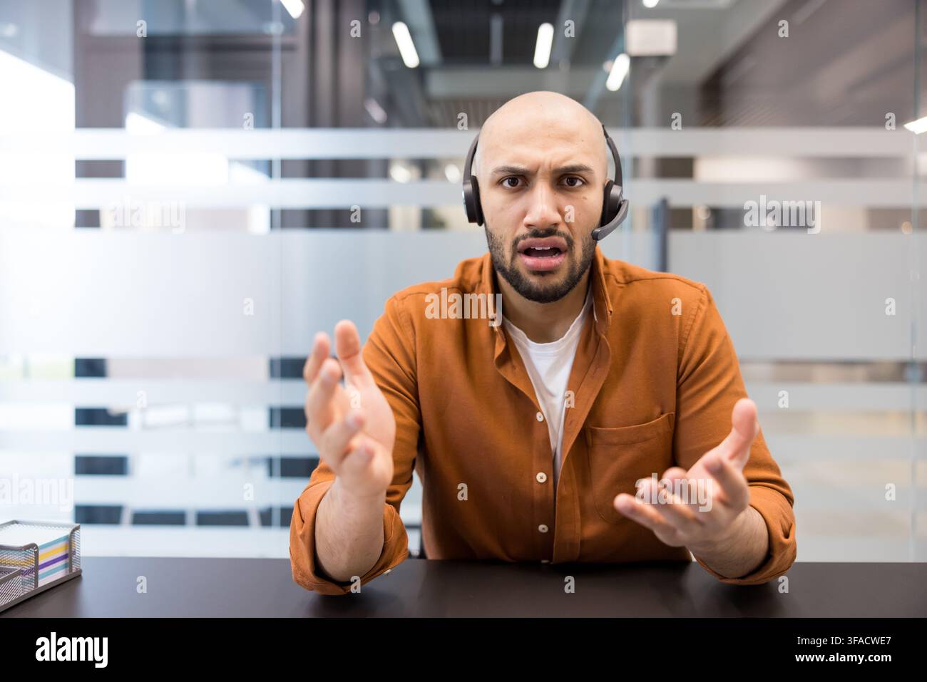Giovane uomo con auricolari sembra stressato e frustrato mentre gesticola attivamente, comunicando un problema o cercando chiarimenti durante un'importante riunione virtuale in un ambiente di ufficio moderno Foto Stock