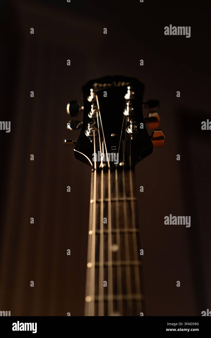 Scatto verticale ad angolo basso di una chitarra elettrica nera che mostra il manico, le corde, i piroli di accordatura e la testa sotto una luce da studio calda e soffusa. Foto Stock