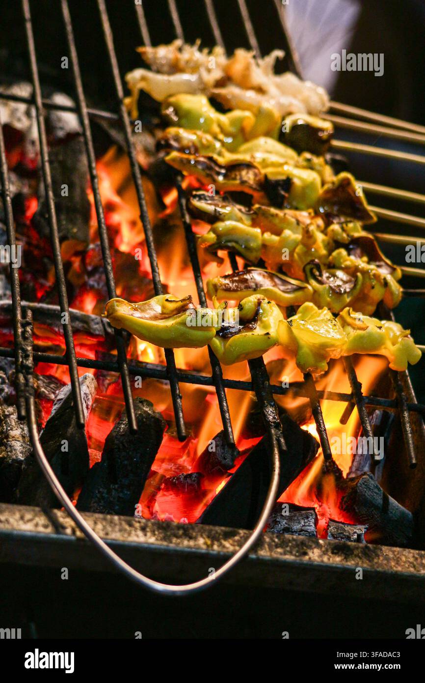 Spiedini di polpo marinati grigliati su fiamme di carbone ad un mercato notturno di cibo di strada thailandese, una scena di cottura di frutti di mare in primo piano. Foto Stock
