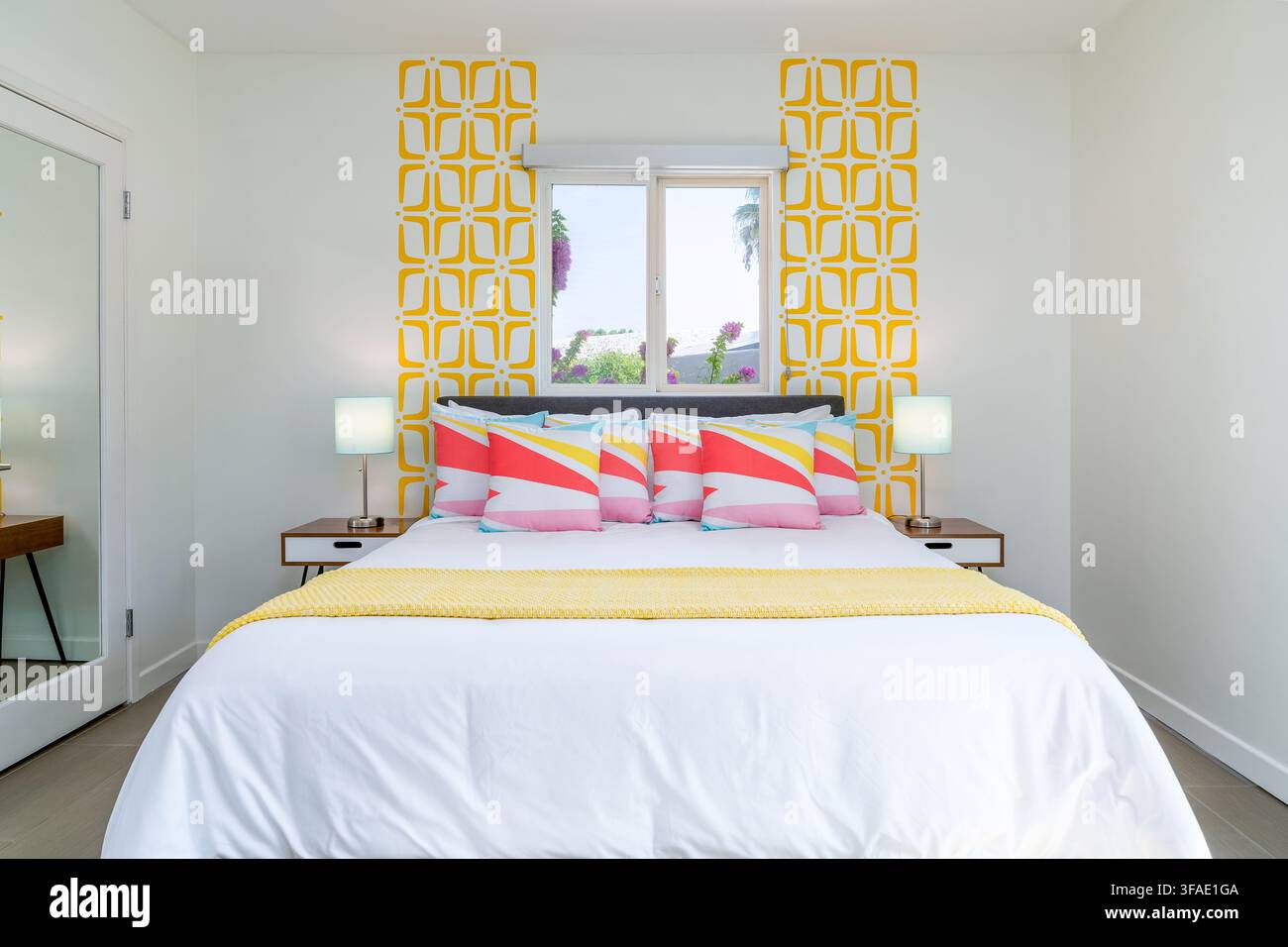 Una camera da letto luminosa e colorata. La stanza presenta un letto bianco con cuscini colorati e una coperta gialla, completata da carta da parati geometrica e na Foto Stock
