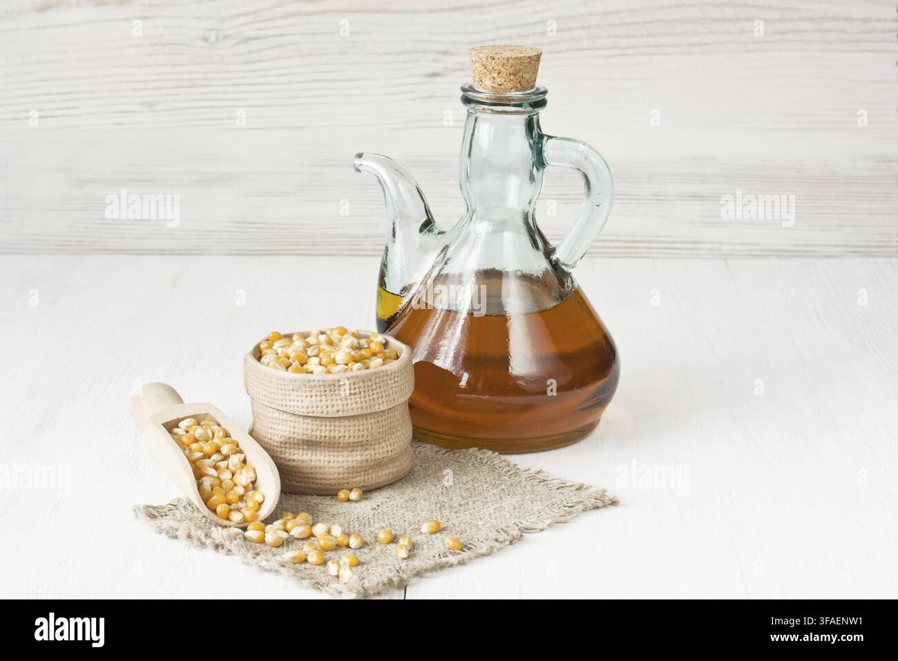 Olio di mais in brocca di vetro e chicchi di mais essiccati su sfondo di legno bianco Foto Stock