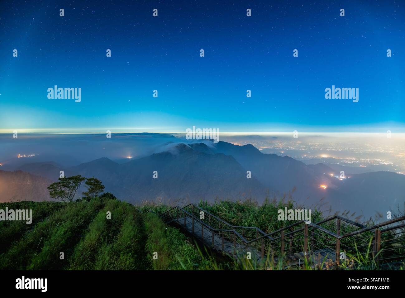 Vista mozzafiato di un cielo notturno stellato che si trasforma in alba sulle catene montuose, nebbia ondulata, una piantagione di tè in primo piano e un villaggio scintillante Foto Stock