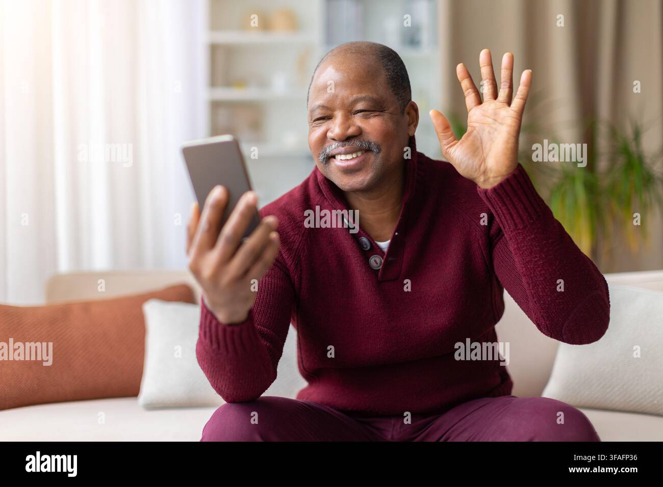 Uomo nero sorridente seduto a casa che saluta durante una videochiamata su smartphone Foto Stock