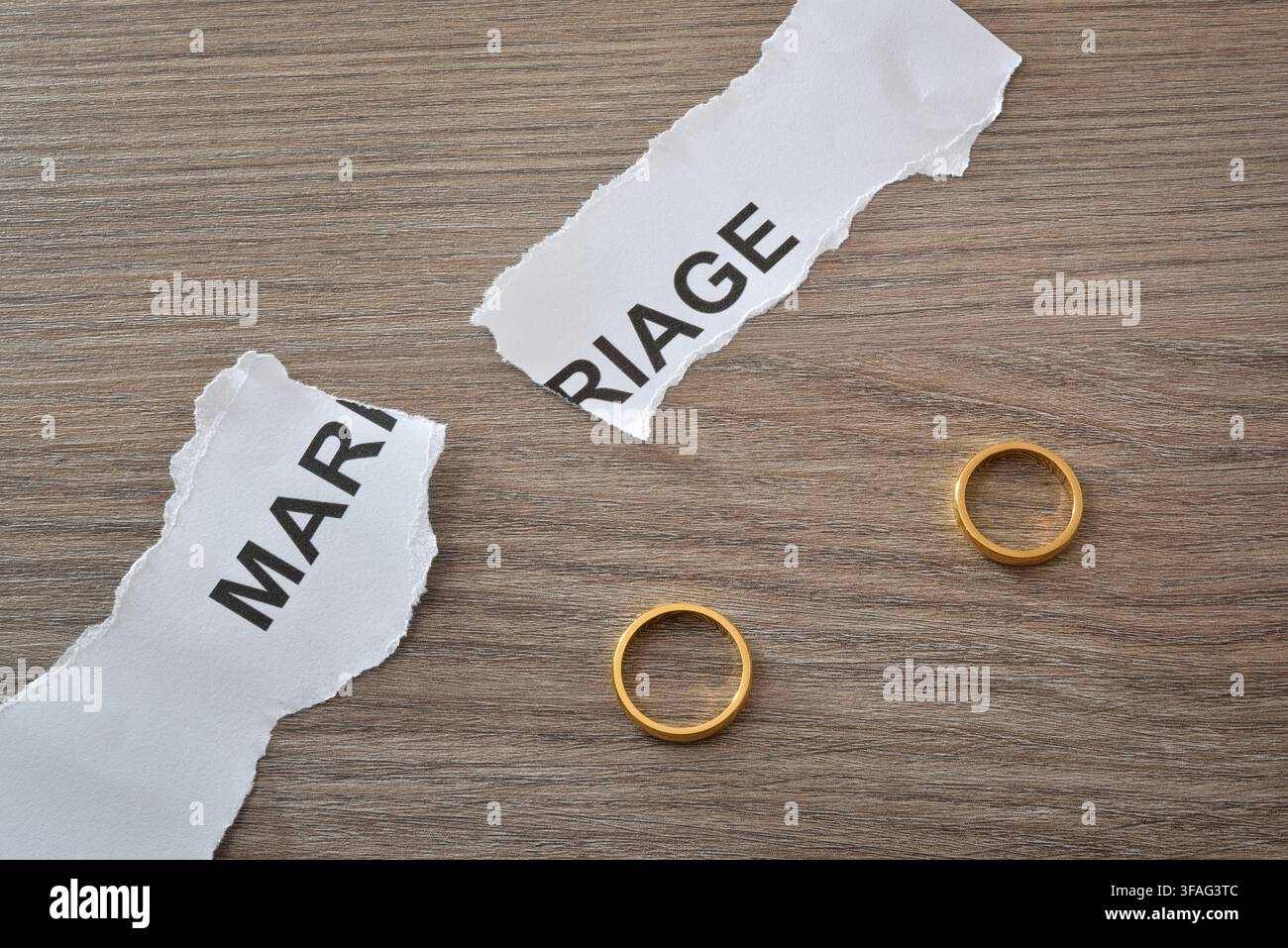 Due anelli nuziali d'oro giacciono separati su una superficie di legno accanto a carta strappata contrassegnata con parole relative al matrimonio, simboleggiando separazione, conflitto e il Foto Stock