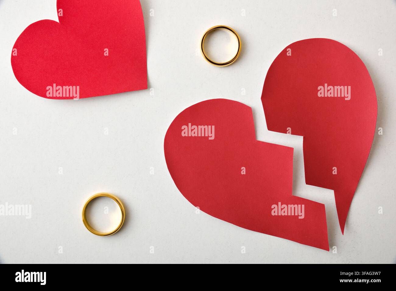 Un cuore di carta rosso rotto giace separato con due anelli d'oro distinti, evocando separazione, perdita, dolore d'amore e la fine di un legame romantico. Foto Stock