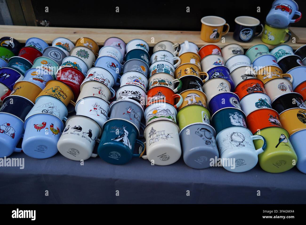 File di tazze smaltate illustrate e colorate in vendita, con vari disegni come paesaggi, animali e testi, su uno stand di mercato affollato. Foto Stock