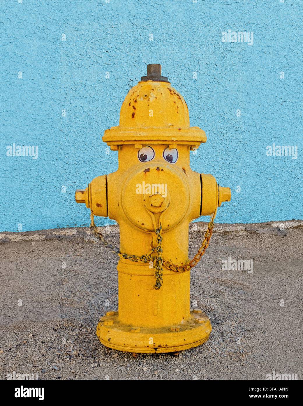 Un allegro idrante giallo con occhi da cartone animato si trova su un muro ruvido. Foto Stock