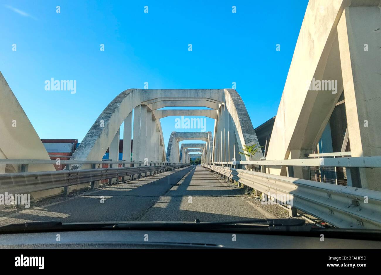 Vista prospettica dall'interno di un'auto che attraversa un moderno ponte ad arco in cemento sotto un cielo blu sereno nell'Italia settentrionale. Foto Stock
