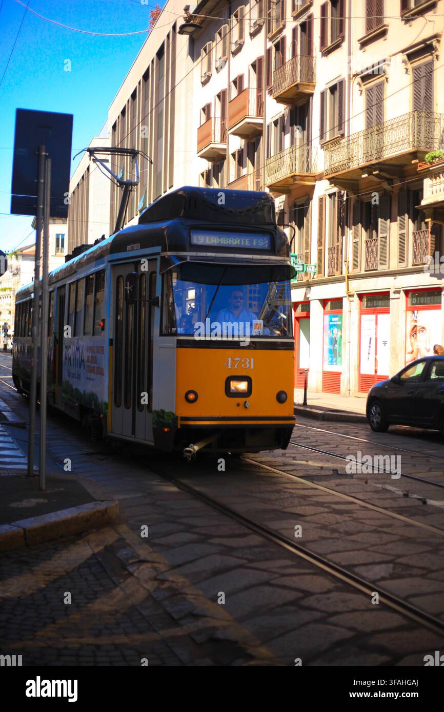 Tram pubblico giallo e bianco numero 19 che procede lungo una strada acciottolata con binari e edifici classici sotto un cielo blu a Milano, Italia. Foto Stock