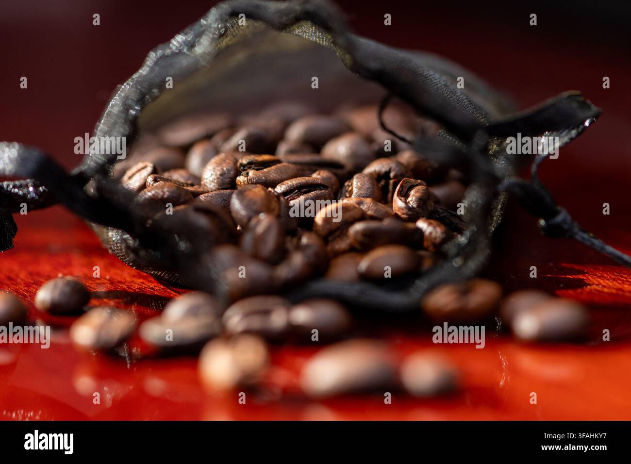 I chicchi di caffè fuoriescono da un piccolo sacchetto a rete nero su una superficie rossa. Foto Stock