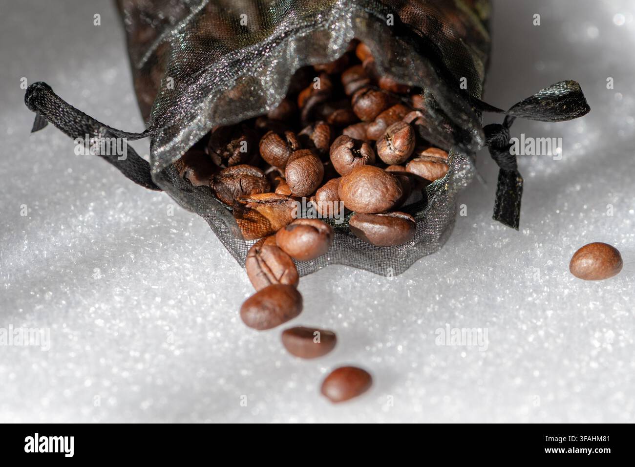 Grani di caffè tostati si spargono da una borsa a rete nera su una superficie bianca. Foto Stock