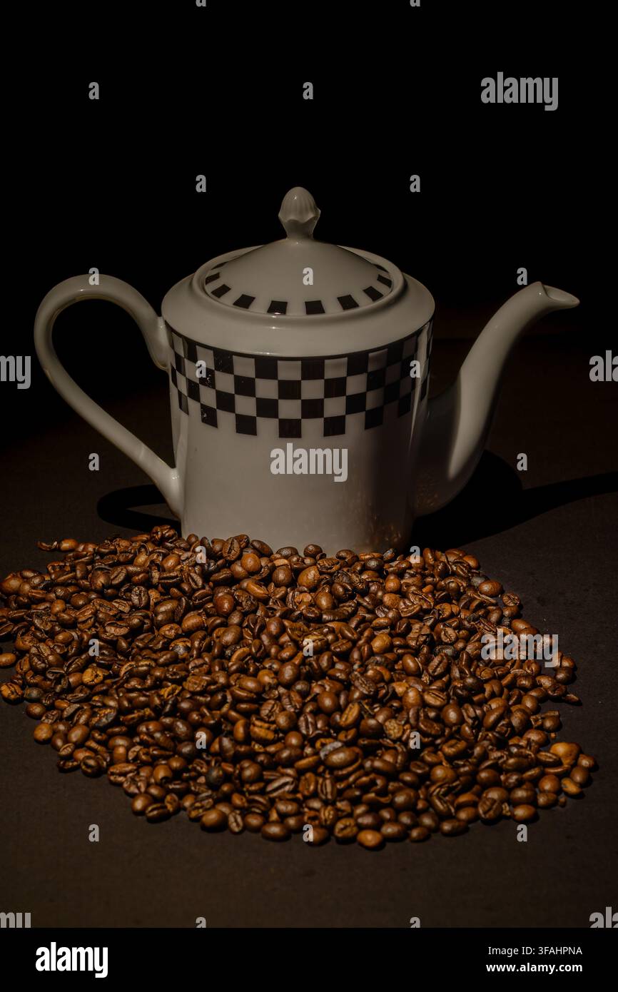 Caffettiera in ceramica bianca a motivi che si trova dietro un grande mucchio di chicchi di caffè tostati su uno sfondo scuro, illuminata da una luce calda e drammatica. Foto Stock