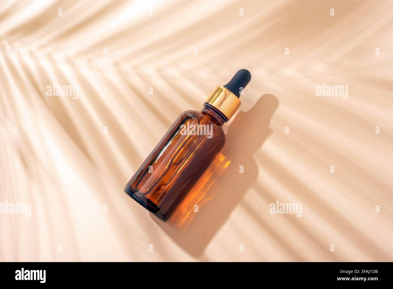 Fiala gocciolatore in ambra illuminata dalla luce calda del sole attraverso una foglia di palma, creando ombre morbide su uno sfondo beige per un'estetica di cura naturale della pelle. Top v Foto Stock