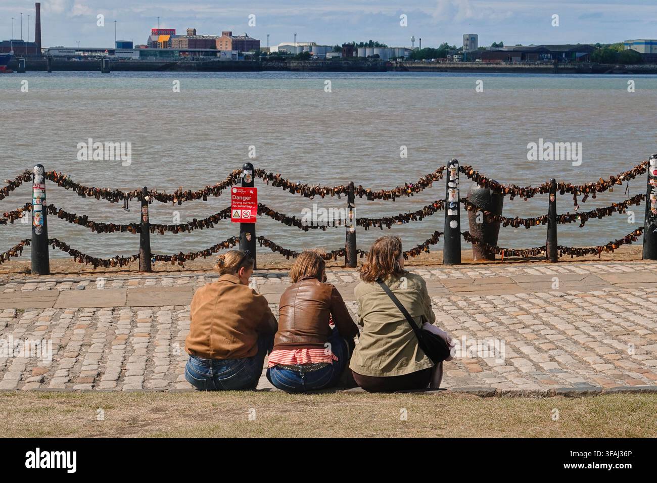 Vista posteriore di tre giovani donne sedute su una passeggiata in ciottoli accanto al fiume Mersey a Liverpool, UK, vicino a ringhiere coperte di lucchetti d'amore Foto Stock