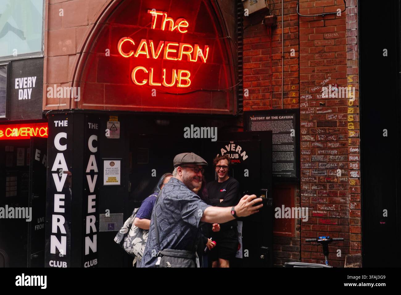Un uomo con un berretto piatto allunga il braccio per scattare un selfie davanti all'insegna al neon iconica all'ingresso del Cavern Club a Liverpool, Regno Unito Foto Stock