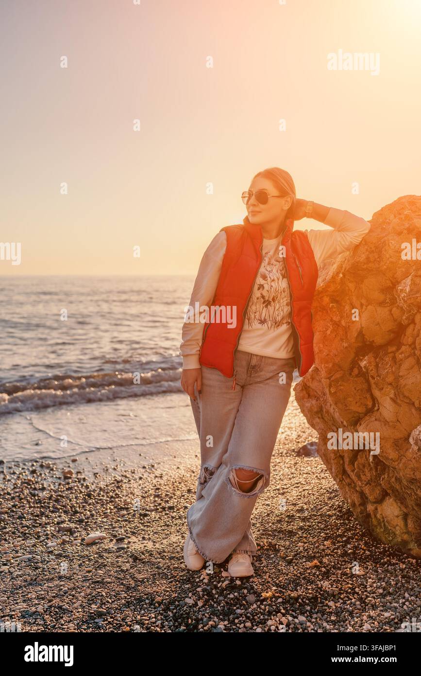 Donna sulla spiaggia al tramonto, appoggiata su una grande roccia durante un'ora dorata serena su una riva di ciottoli con spazio di copia Foto Stock