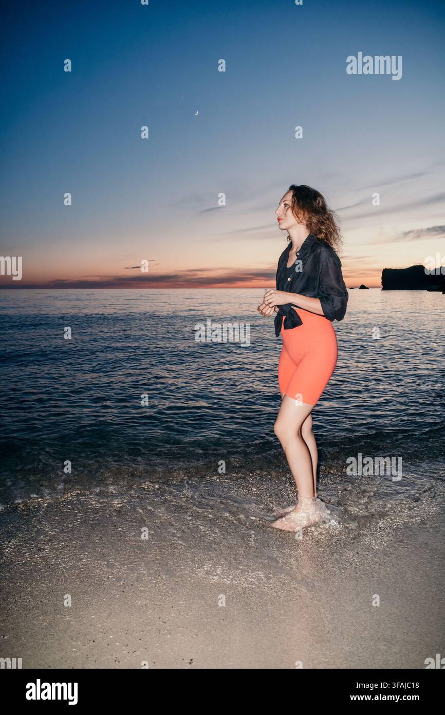 Donna serena in piedi sulla riva dell'oceano durante il crepuscolo, godendosi momenti tranquilli all'aperto Foto Stock
