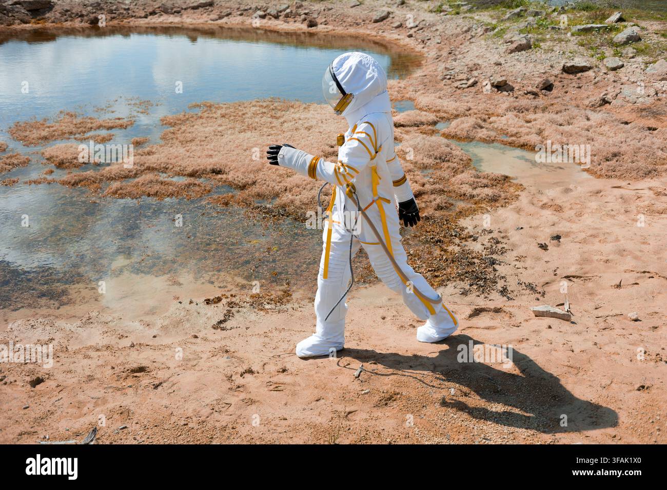 Esploratrice femminile in una tuta spaziale retrò bianca e gialla che cammina accanto a un lago poco profondo su terreno sabbioso rosso, un concetto di ricerca sugli esopianeti e la ricerca f Foto Stock