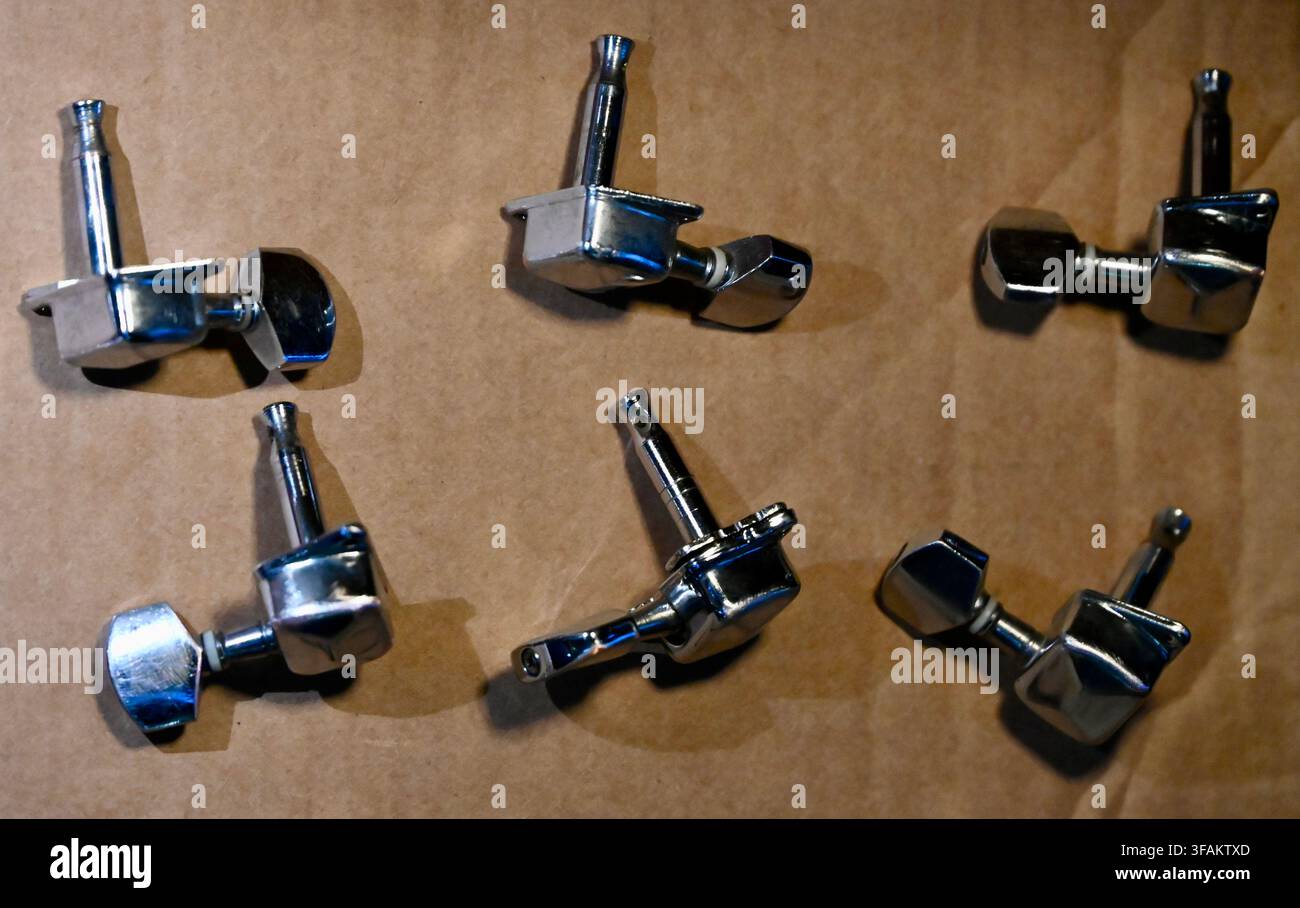 Una fotografia commerciale dall'alto di un set completo di sei meccanismi di accordatura per chitarra elettrica cromati lucidi (teste di macchina) disposti ordinatamente su una superficie marrone ruvida. La finitura metallica lucida e i dettagli degli ingranaggi meccanici sono chiaramente visibili, evidenziando l'hardware e le parti degli strumenti musicali. Foto Stock