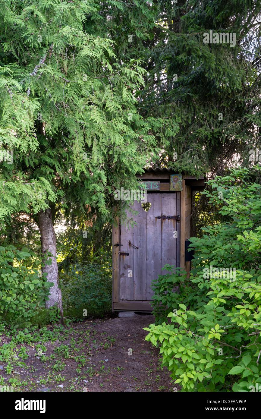 Toilette esterna in legno rustico con un'apertura a forma di cuore nella porta, circondata da fitta vegetazione verde e alberi. Foto Stock