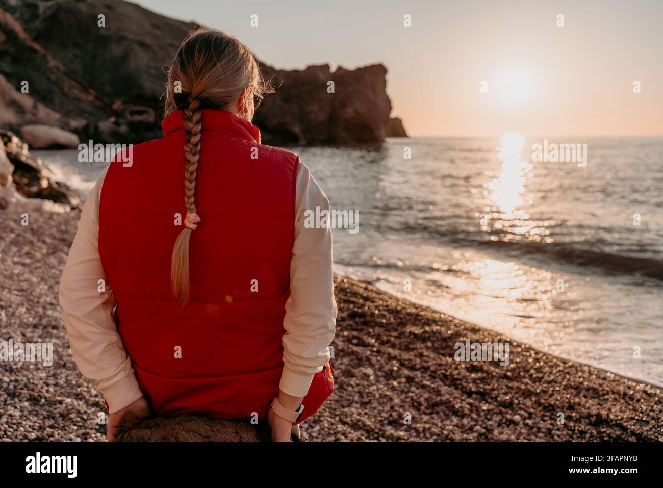 Donna del tramonto donna sulla spiaggia con un giubbotto rosso si siede su una riva rocciosa guardando il sole che tramonta sul mare Foto Stock