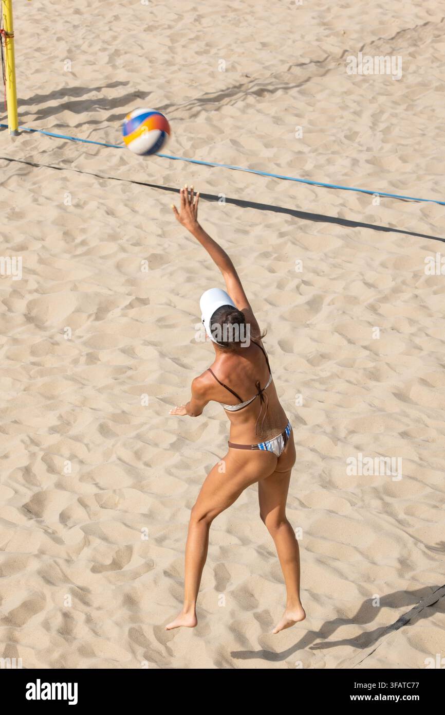 Vista posteriore ad angolazione alta di una donna atletica in bikini che si allunga per servire una palla da volley durante una partita su un campo da spiaggia sabbioso. Foto Stock