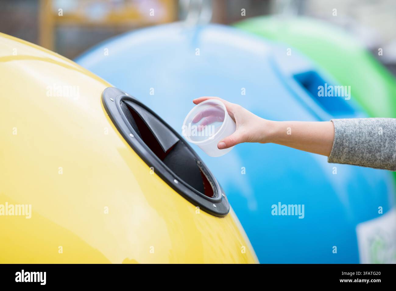Mano irriconoscibile che deposita un contenitore di plastica in un bidone del riciclo colorato, gestione dei rifiuti all'aperto Foto Stock