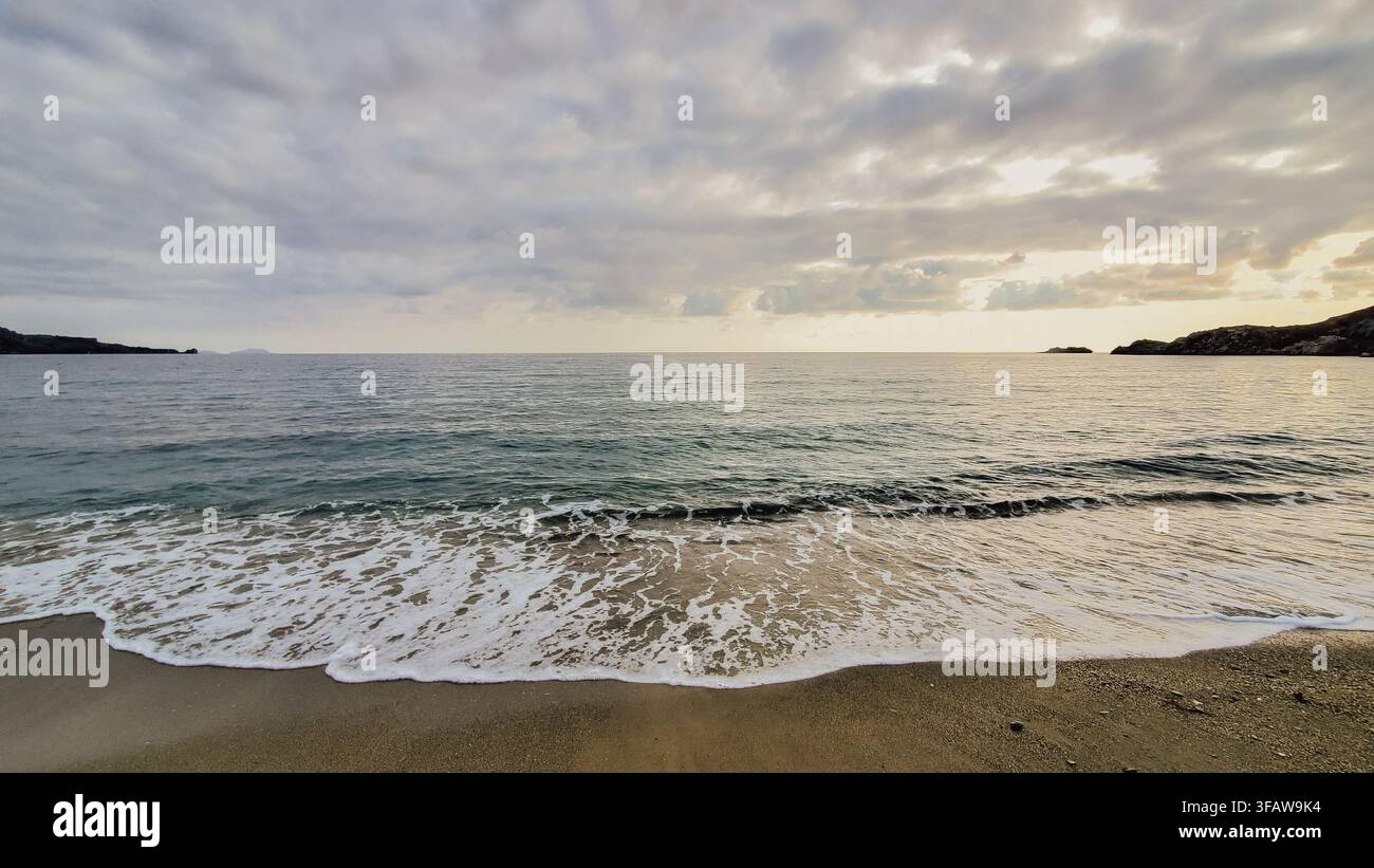Onde del mare che schiumano e lavano la spiaggia sabbiosa sotto un cielo nuvoloso durante il tramonto sulla costa mediterranea della Grecia foto d’archivio. Foto Stock