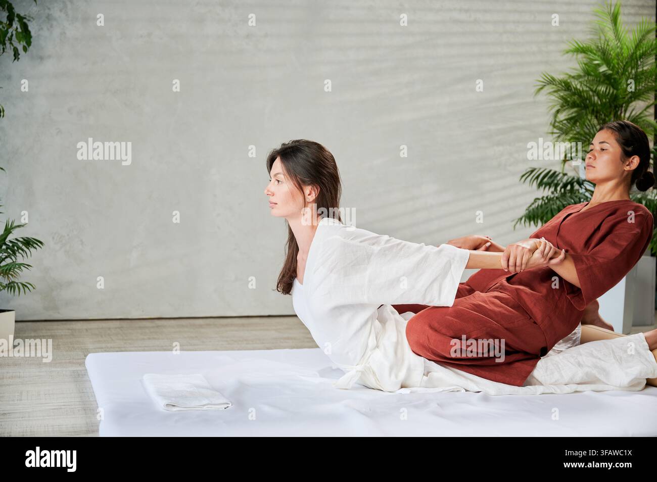 Massaggiatrice che fa un massaggio yoga tailandese. Terapeuta donna che fa un massaggio tradizionale, allungando il corpo di una paziente. I clienti sono sdraiati sul tappetino e si godono un massaggio alle mani in un'atmosfera tranquilla. Foto Stock