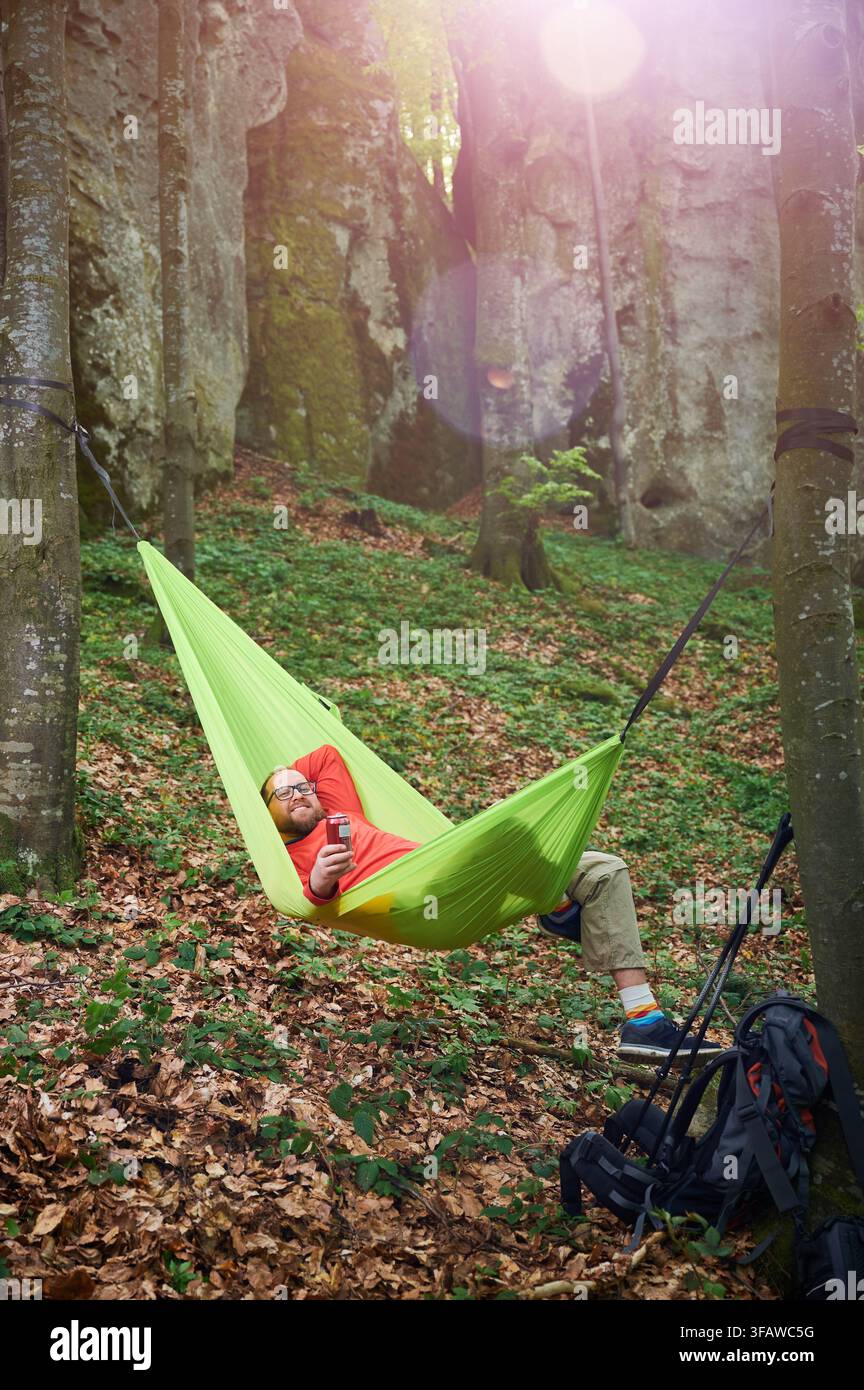 Persona che si rilassa in un'amaca verde brillante legata tra due alberi in una lussureggiante foresta verde vicino alle rocce. L'uomo turista tiene un drink, godendo di momenti tranquilli circondati dalla natura. Zaino e attrezzatura nelle vicinanze. Foto Stock