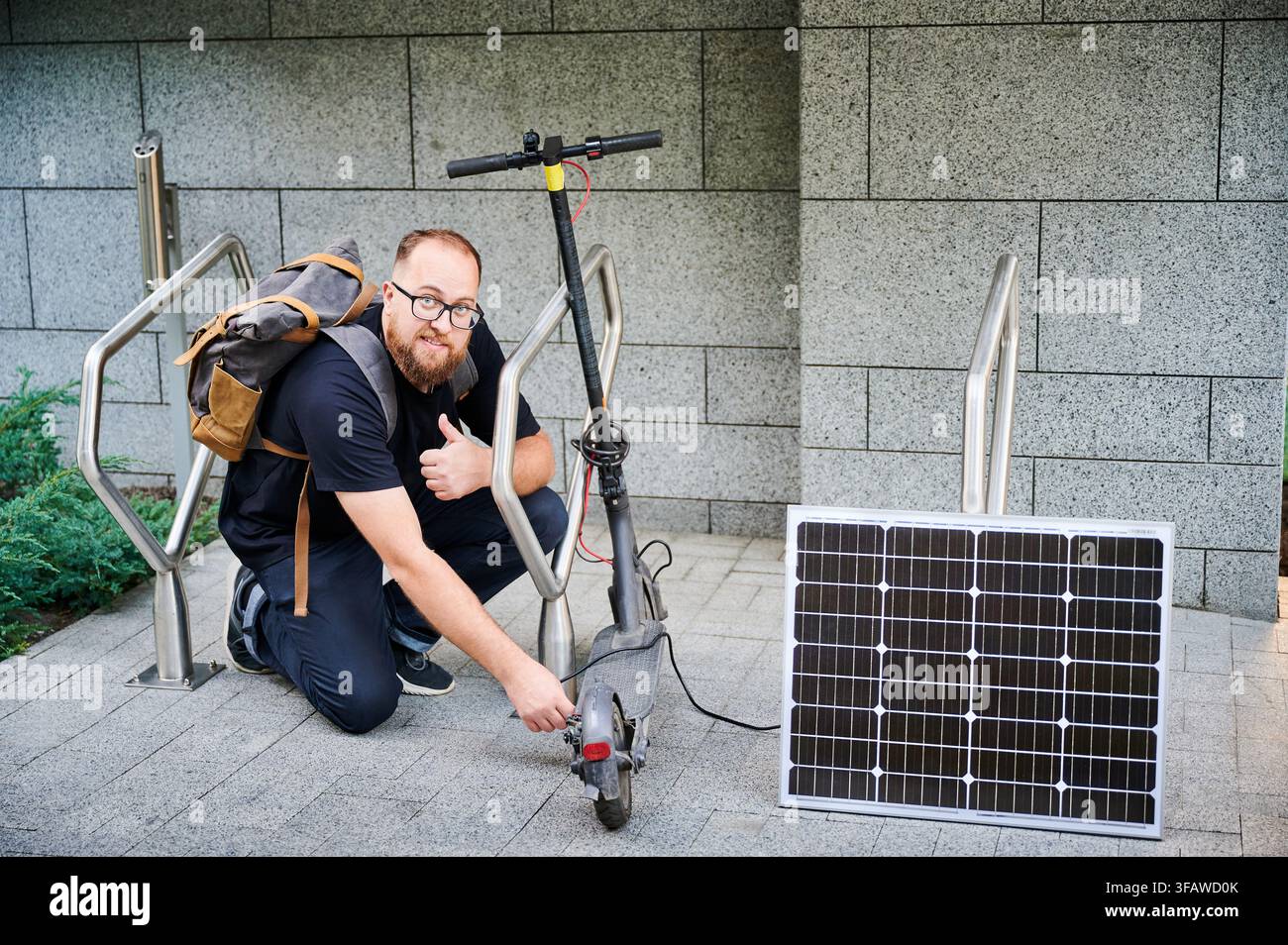 Uomo collega un pannello solare a uno scooter elettrico per caricarlo, dando il pollice alzato. Applicazione pratica dell'energia rinnovabile sostenibile. Concetto di integrazione della tecnologia ecologica nei trasporti. Foto Stock
