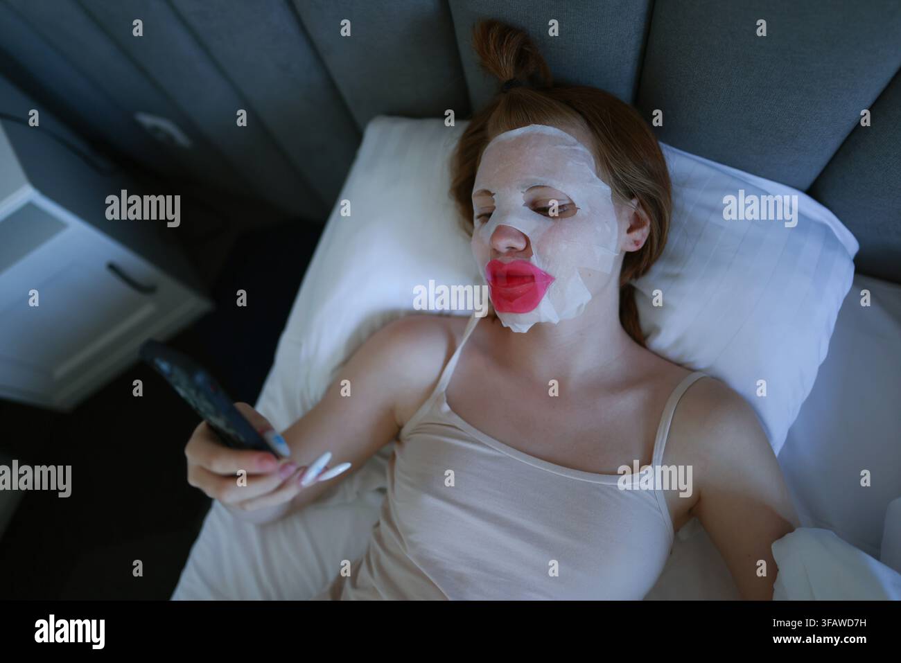 Giovane donna che si rilassa a letto con una maschera in foglio e un patch per le labbra mentre utilizza lo smartphone durante la sua routine di cura della pelle a casa. Foto Stock