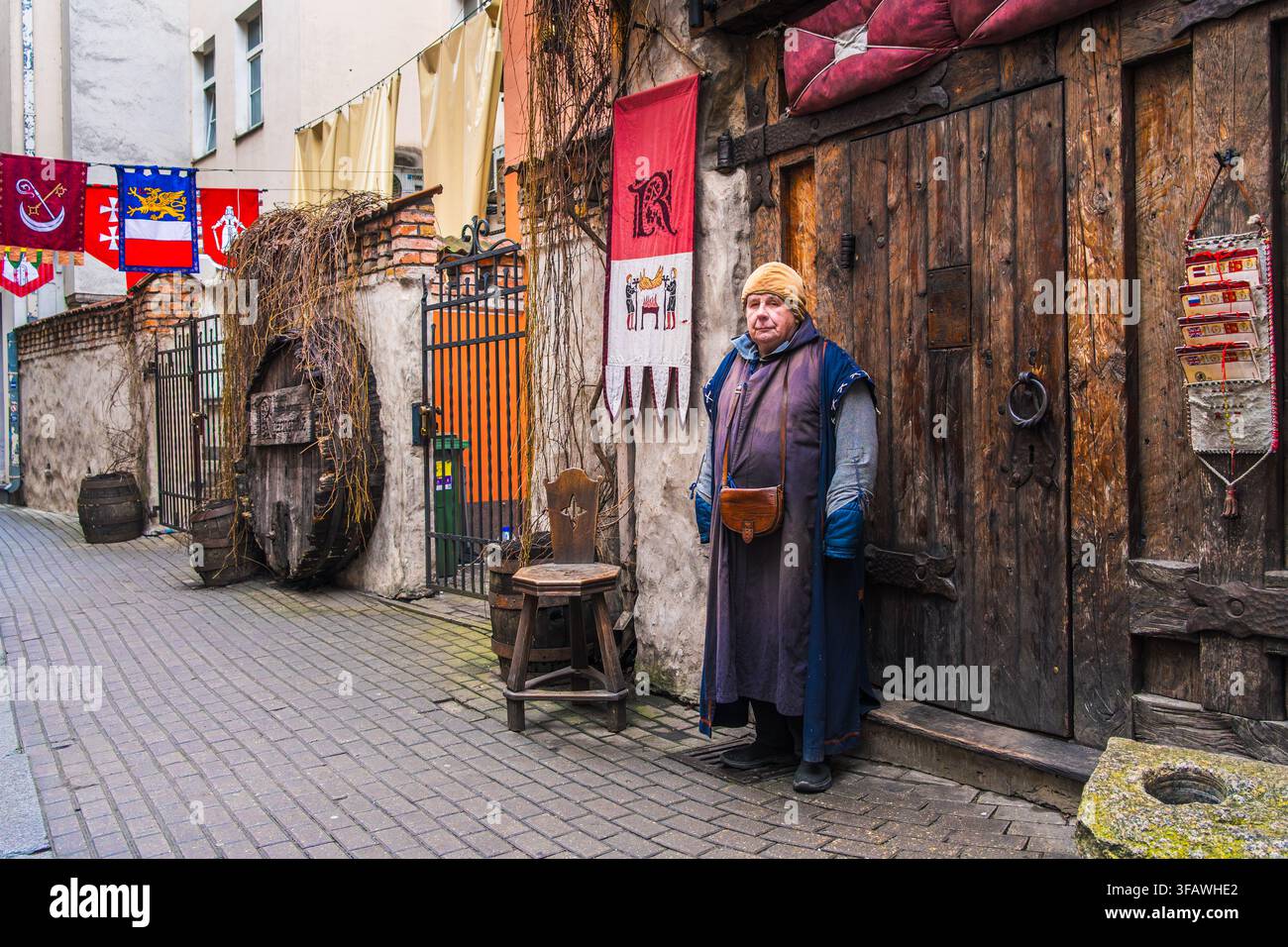 Un incontro con la storia vivente appare mentre un uomo in costume di mezza età si trova accanto a porte di legno e cartelli artigianali in un vicolo della vecchia città per il turismo culturale. Riga Foto Stock